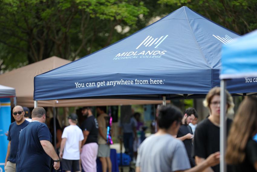 In focus a blue tent with MTC logo and tagline "You can get anywhere from here." Attendees of the Summer 2024 Open House are out of focus in the back and foreground. 