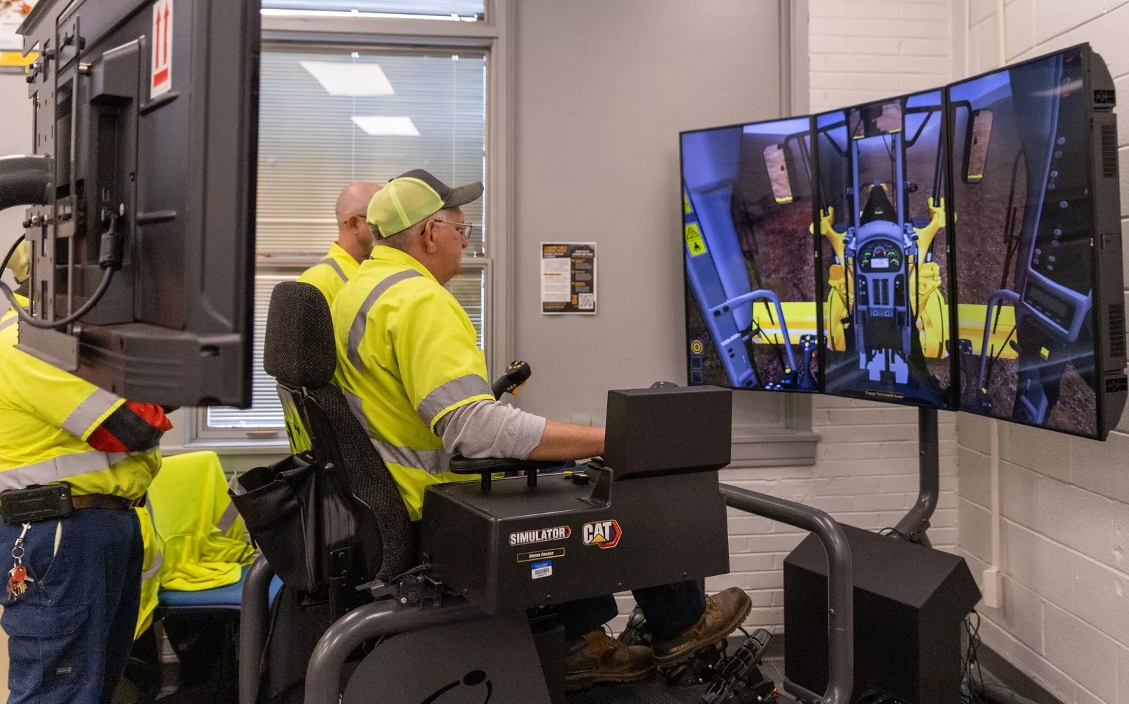 Two individuals in high-visibility vests operate a CAT heavy equipment simulator with a three-screen display in a training facility.